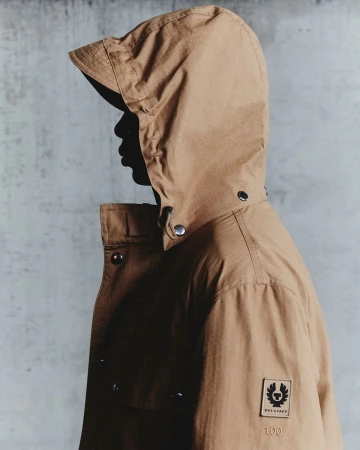 Profile view of a person wearing a camel hooded jacket against a textured concrete wall, photographed in London.