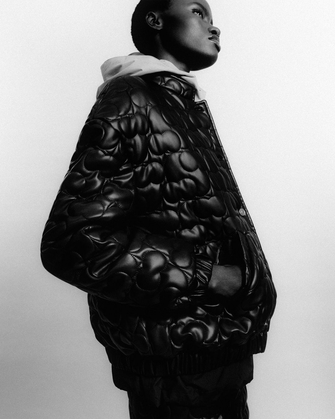 Monochrome fashion portrait of a Black female model in a dark quilted jacket with a light hoodie against a plain white background.