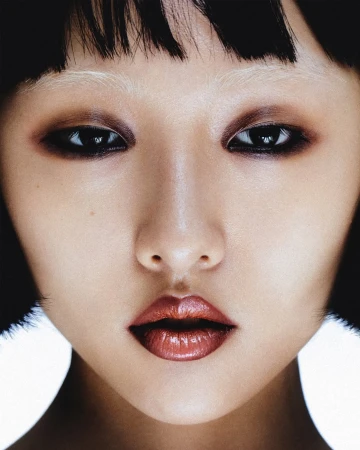 Close-up portrait of a young Asian woman with dark eyes, glossy lips, short black hair and bleached eyebrows in a studio setting.