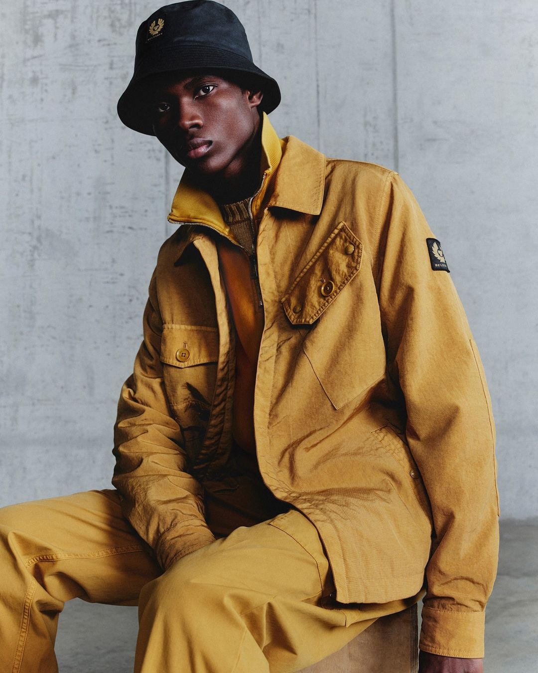 Portrait of a young Black man in a yellow jacket and bucket hat sitting indoors against a concrete wall, fashion editorial style.