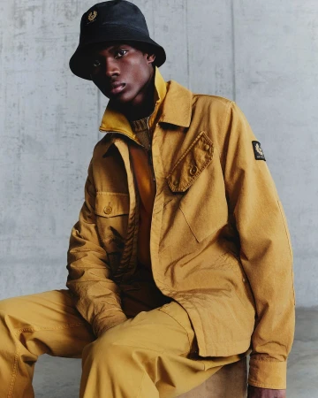 Portrait of a young Black man in a yellow jacket and bucket hat sitting indoors against a concrete wall, fashion editorial style.