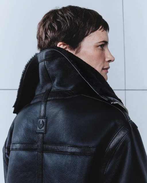 Adult woman with short brown hair poses in a black leather shearling coat against a light grey background in a studio shot.