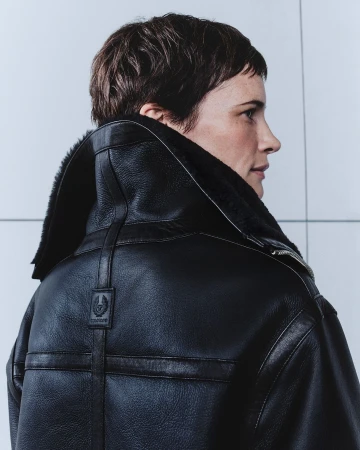 Adult woman with short brown hair poses in a black leather shearling coat against a light grey background in a studio shot.