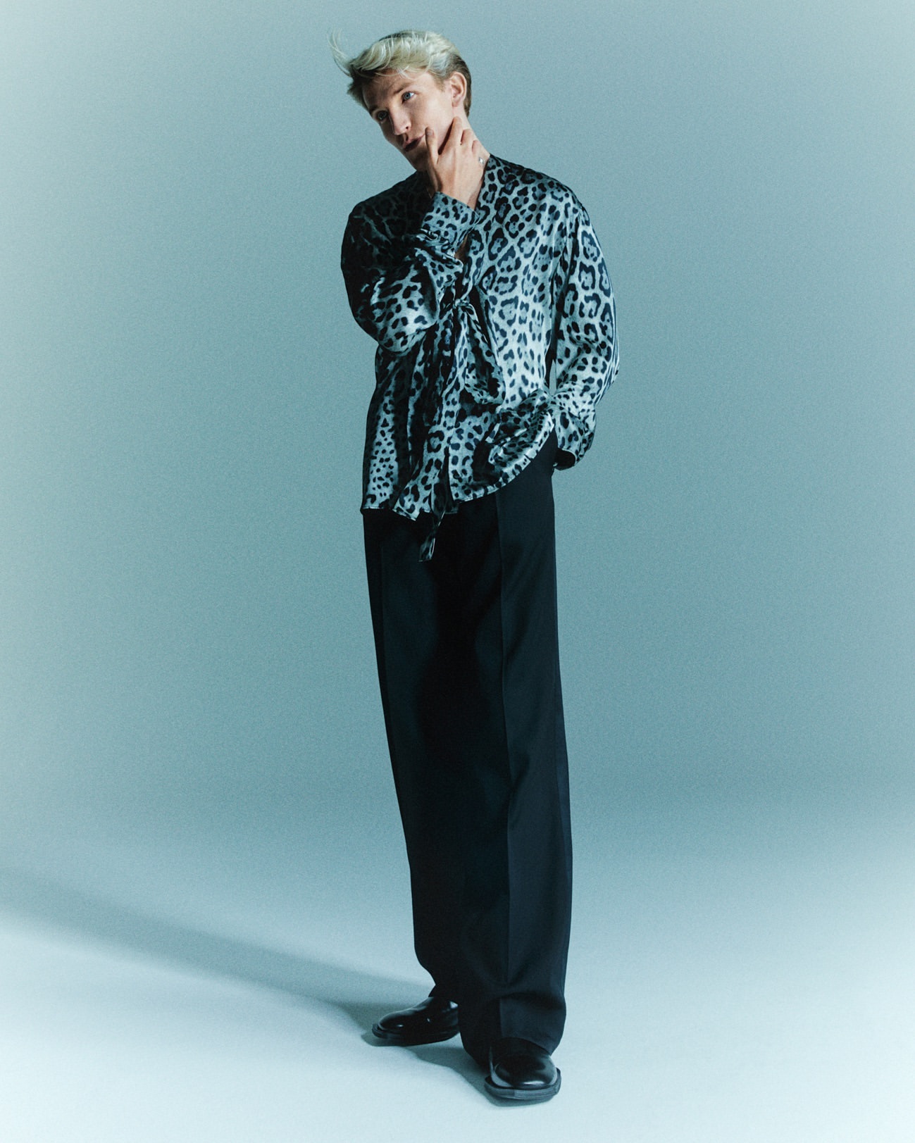 A fashionable, light-haired man in a leopard-print shirt poses in a studio, wearing black trousers and dress shoes against a light blue backdrop.