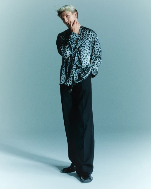 A fashionable, light-haired man in a leopard-print shirt poses in a studio, wearing black trousers and dress shoes against a light blue backdrop.