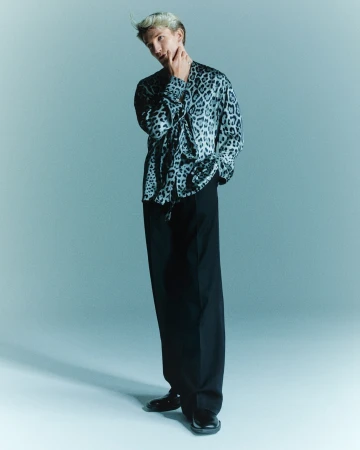 A fashionable, light-haired man in a leopard-print shirt poses in a studio, wearing black trousers and dress shoes against a light blue backdrop.
