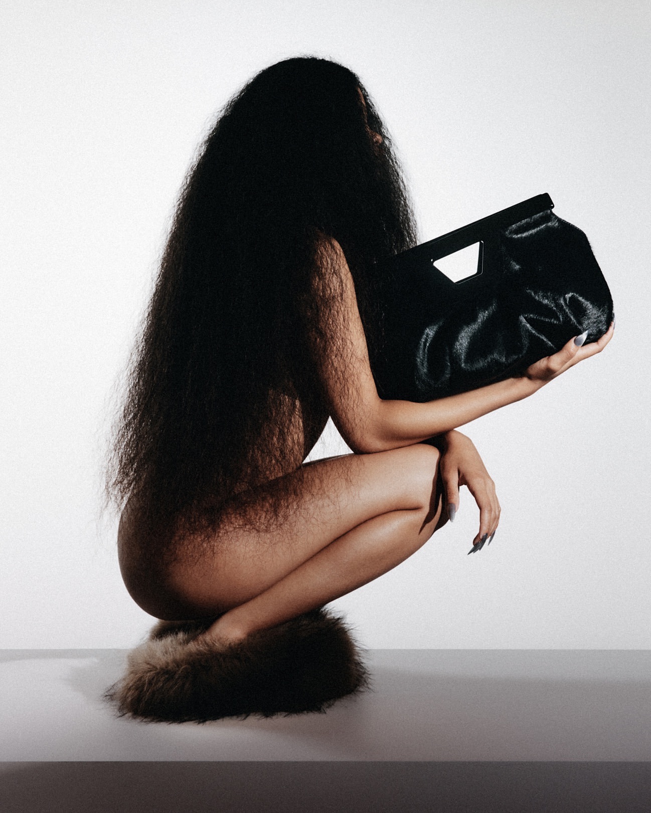 Woman with long dark hair crouches and holds a large black furry bag in a studio setting against a white background. Fashion.