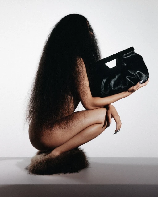 Woman with long dark hair crouches and holds a large black furry bag in a studio setting against a white background. Fashion.