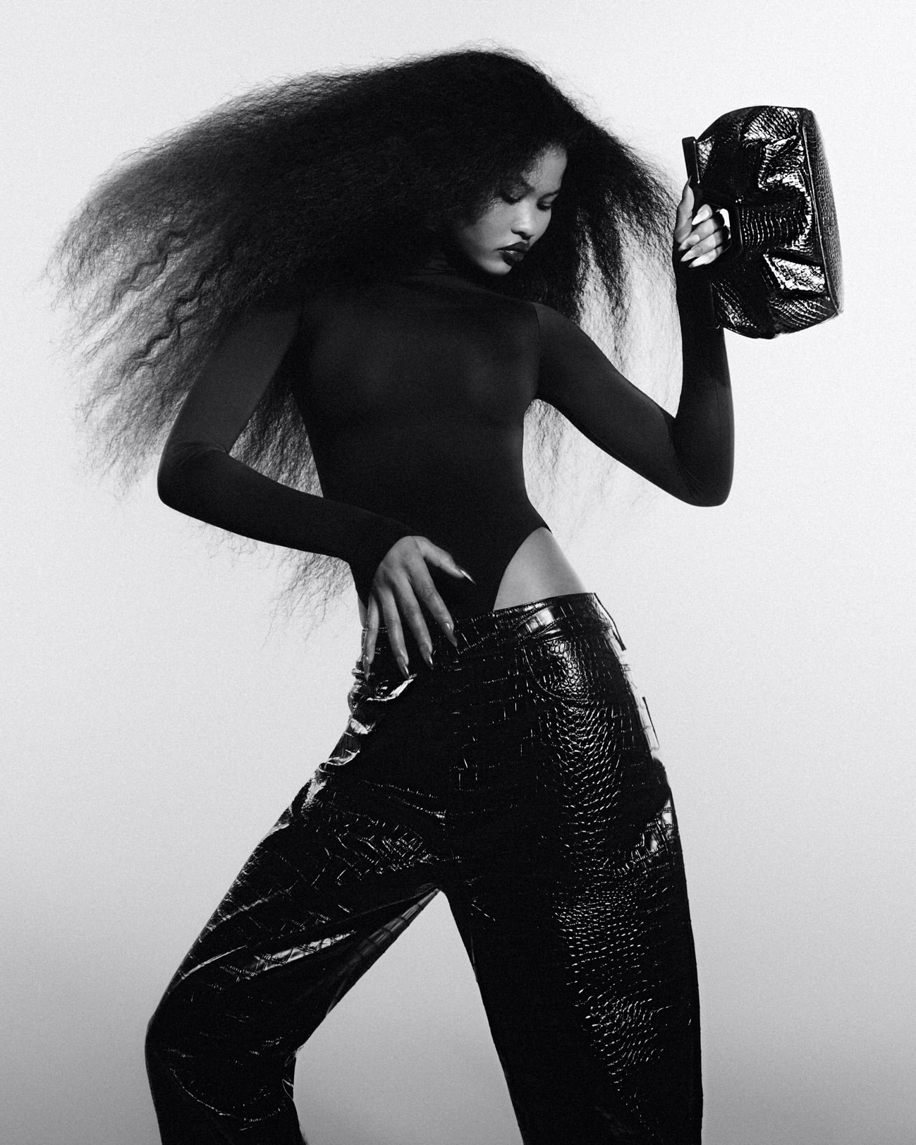 A person with afro hairstyle is posing in a dark bodysuit and wide black faux crocodile pants, holding a shiny black clutch in a photo studio.