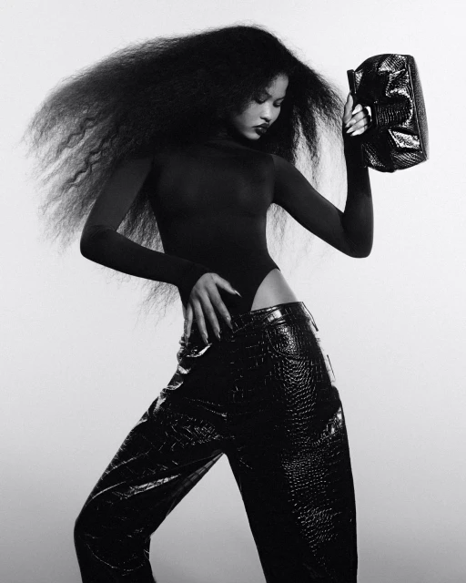 A person with afro hairstyle is posing in a dark bodysuit and wide black faux crocodile pants, holding a shiny black clutch in a photo studio.