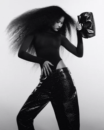 A person with afro hairstyle is posing in a dark bodysuit and wide black faux crocodile pants, holding a shiny black clutch in a photo studio.
