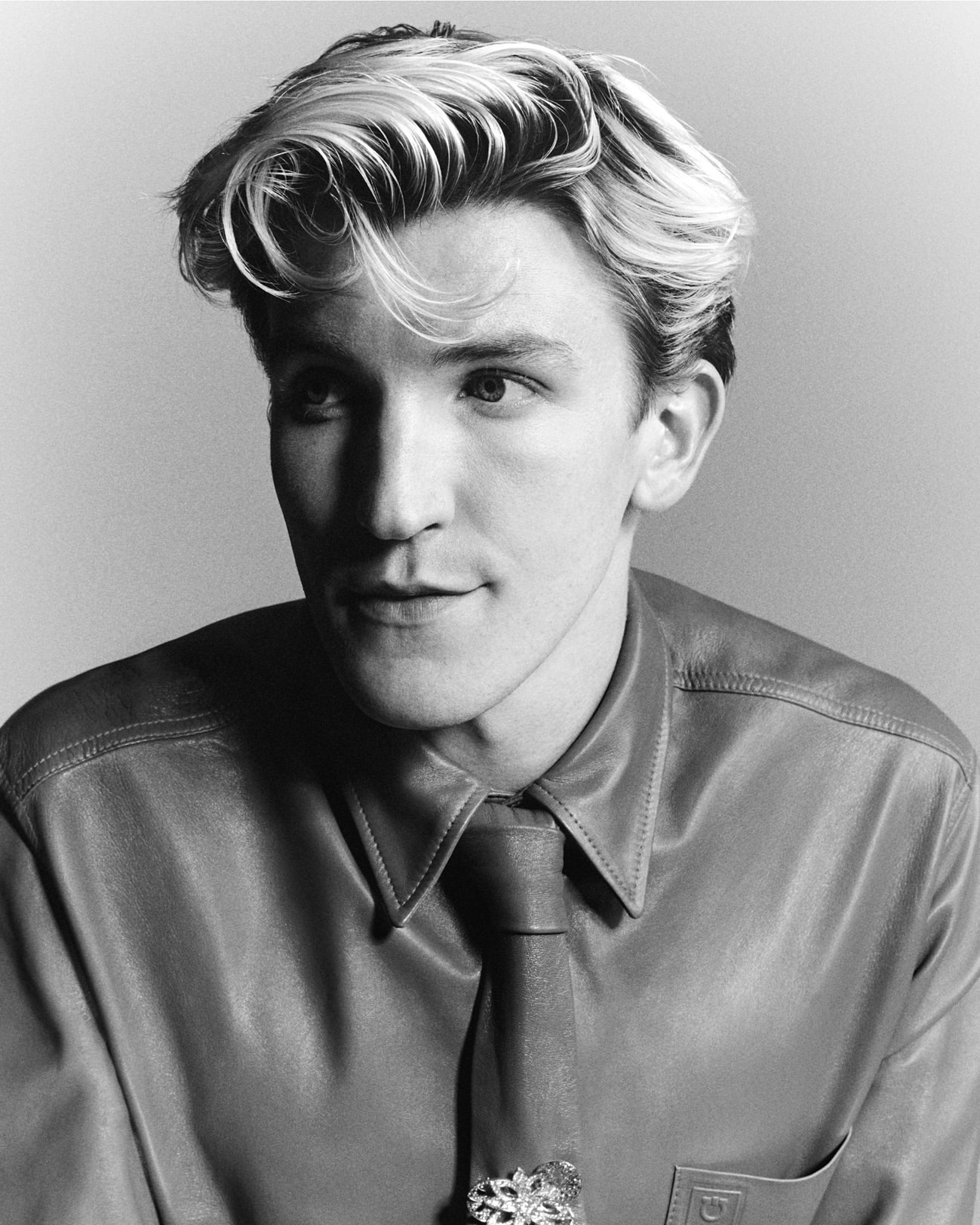 Black and white portrait of an adult male with blond-streaked hair wearing a shirt and tie with a decorative pin. Neutral background.