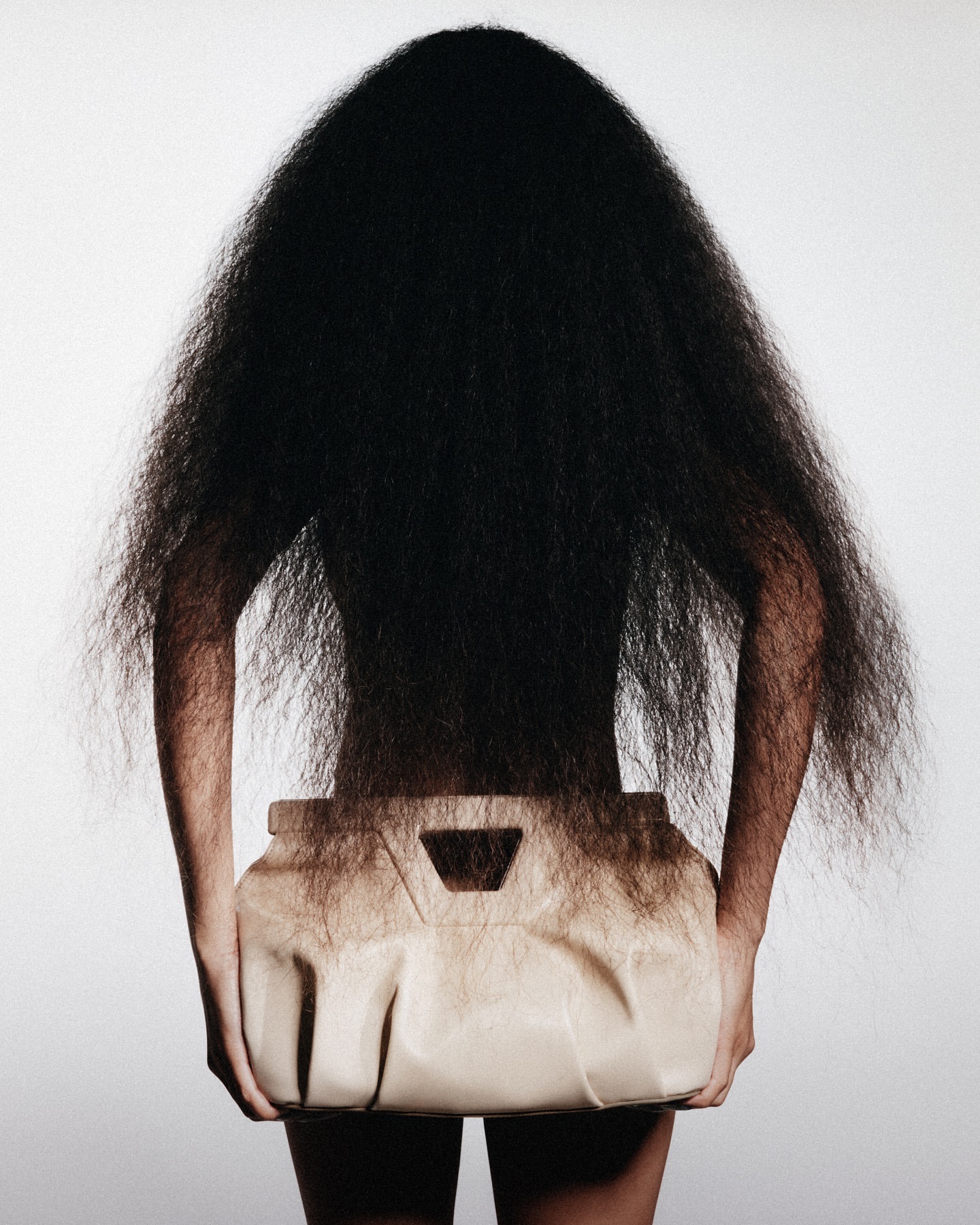 Back view of model with an exaggerated, teased hairstyle, holding a light tan leather handbag, set against a light grey backdrop.