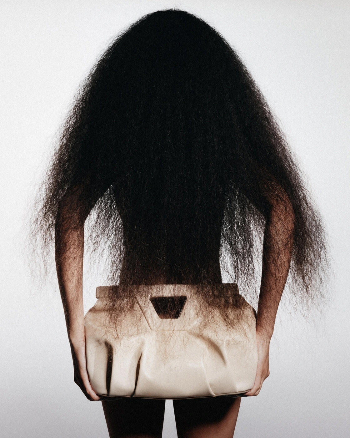 Back view of a person with long dark voluminous hair holding a structured beige leather handbag against white background