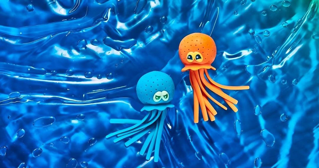 Two animated toy jellyfish, one orange and one blue, sit on a vibrant blue liquid surface, creating a playful and whimsical scene.