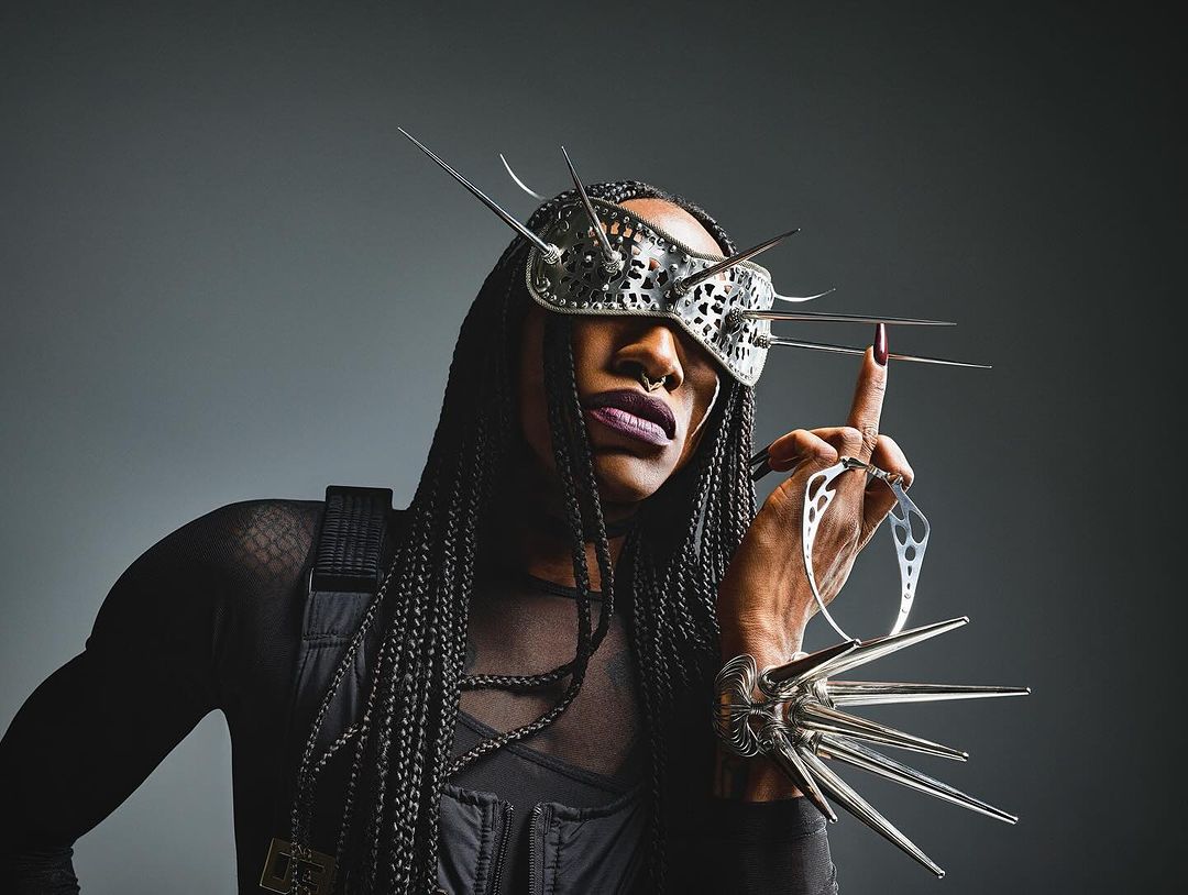 Person wearing a futuristic metal mask with spikes and unique jewelry, posing against a gray backdrop in a studio setting.