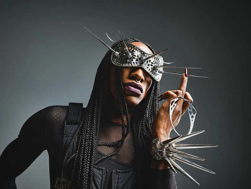 Person wearing a futuristic metal mask with spikes and unique jewelry, posing against a gray backdrop in a studio setting.