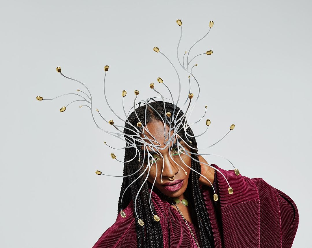 A woman with braided hair and a dramatic silver and gold headpiece poses against a light grey background, wearing a maroon pleated top.