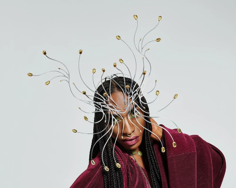 A woman with braided hair and a dramatic silver and gold headpiece poses against a light grey background, wearing a maroon pleated top.