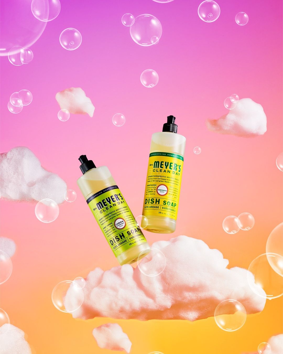 Two bottles of Mrs. Meyer's Clean Day dish soap float among white clouds and bubbles against a pink and orange gradient background.