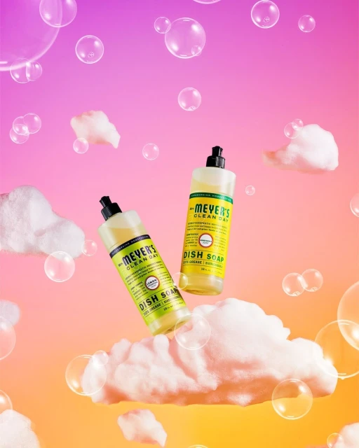 Two bottles of Mrs. Meyer's Clean Day dish soap float among white clouds and bubbles against a pink and orange gradient background.