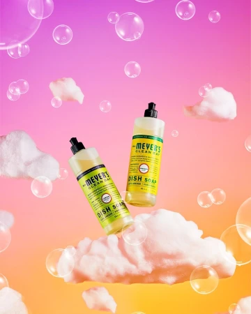 Two bottles of Mrs. Meyer's Clean Day dish soap float among white clouds and bubbles against a pink and orange gradient background.