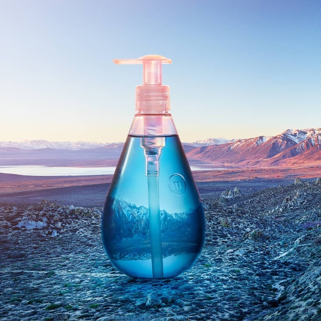 Product shot of a clear plastic bottle with blue liquid and pump dispenser against a scenic mountain and lake landscape backdrop.