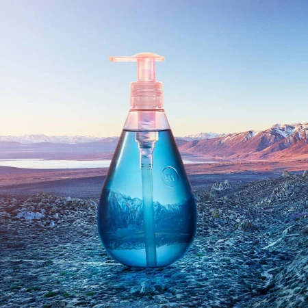 Product shot of a clear plastic bottle with blue liquid and pump dispenser against a scenic mountain and lake landscape backdrop.