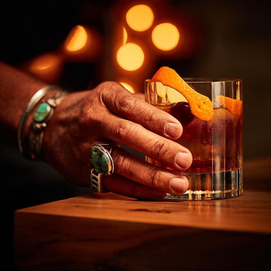 Close-up of a hand holding a glass of a dark cocktail garnished with an orange peel twist, showcasing jewelry and a warm ambiance.