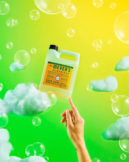 Mrs. Meyer's Clean Day hand soap refill floats above a finger surrounded by soap bubbles against a graduated yellow-green background.