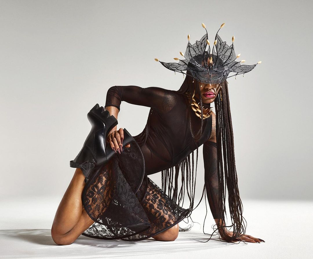 A person with long braids poses in a sheer black dress and elaborate headpiece, kneeling on the floor with one leg holding a black boot.