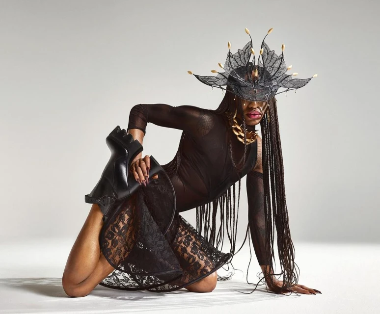 A person with long braids poses in a sheer black dress and elaborate headpiece, kneeling on the floor with one leg holding a black boot.