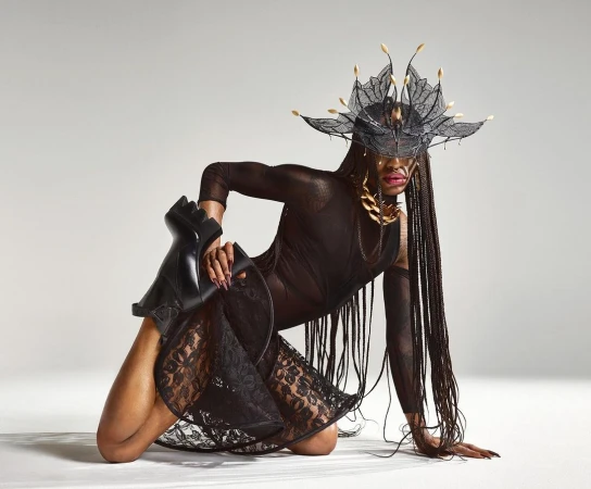 A person with long braids poses in a sheer black dress and elaborate headpiece, kneeling on the floor with one leg holding a black boot.