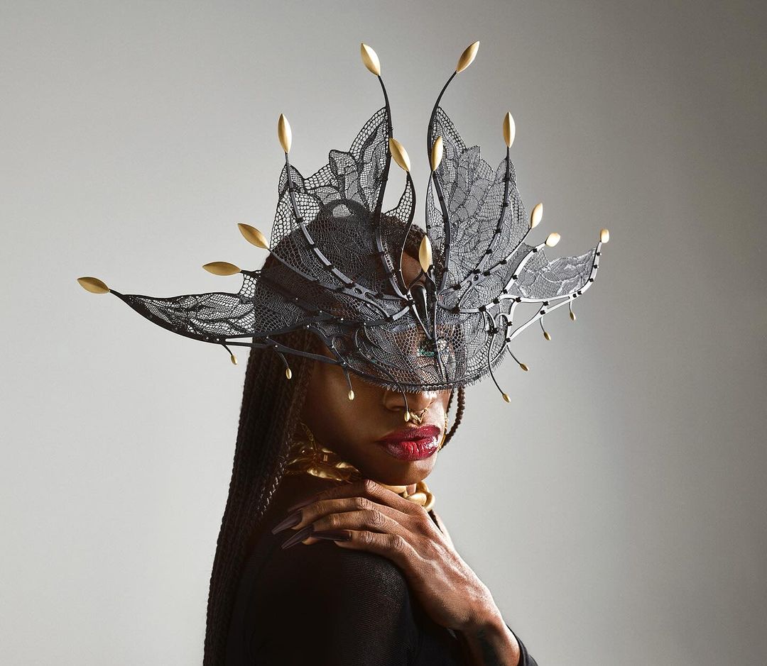 Close-up portrait of a Black woman with an elaborate black lace and gold headdress, gold collar, and long braided hair against a grey backdrop.