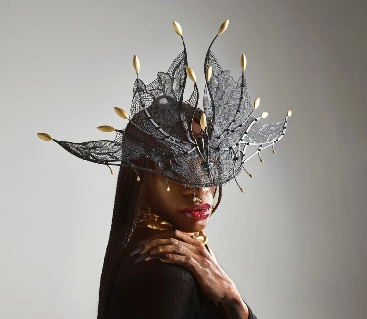 Close-up portrait of a Black woman with an elaborate black lace and gold headdress, gold collar, and long braided hair against a grey backdrop.