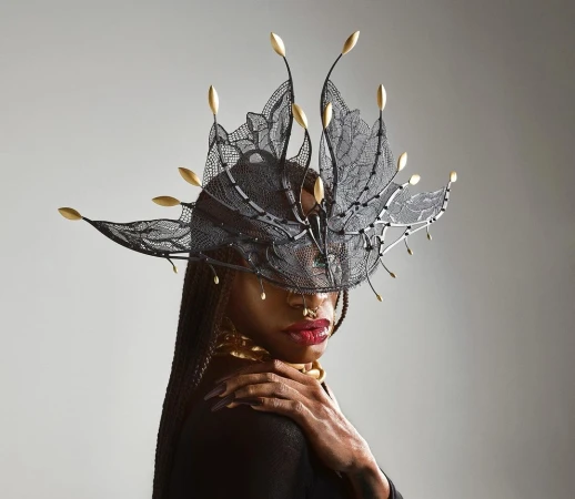 Close-up portrait of a Black woman with an elaborate black lace and gold headdress, gold collar, and long braided hair against a grey backdrop.
