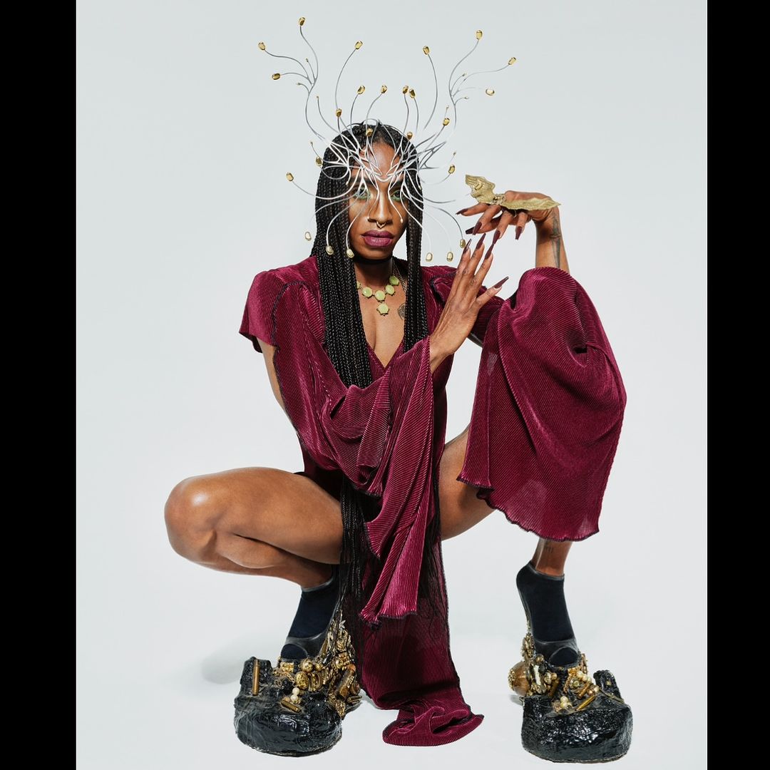 Black model crouching in a burgundy textured outfit, wearing an intricate wire headdress and sculptural platform shoes against a white background.