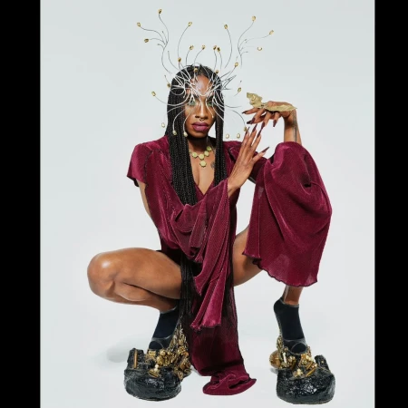 Black model crouching in a burgundy textured outfit, wearing an intricate wire headdress and sculptural platform shoes against a white background.