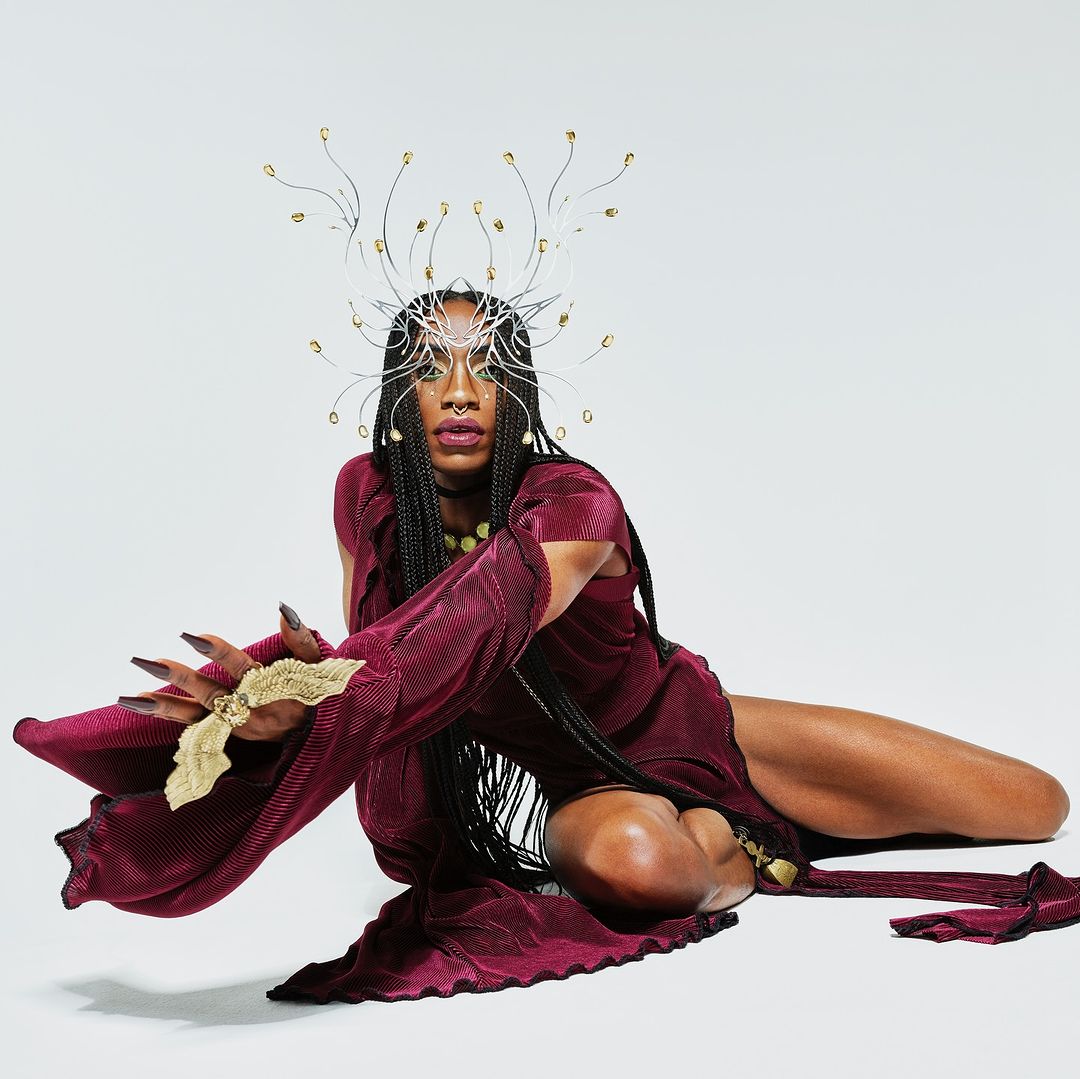 Person wearing a burgundy dress and elaborate headpiece sits on a white background. The image showcases beauty and fashion elements.