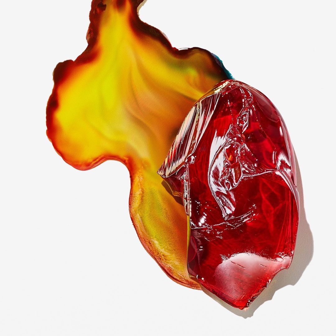 Abstract still life photography featuring a red translucent geometric shape and a yellow-orange fluid shape on a white background.