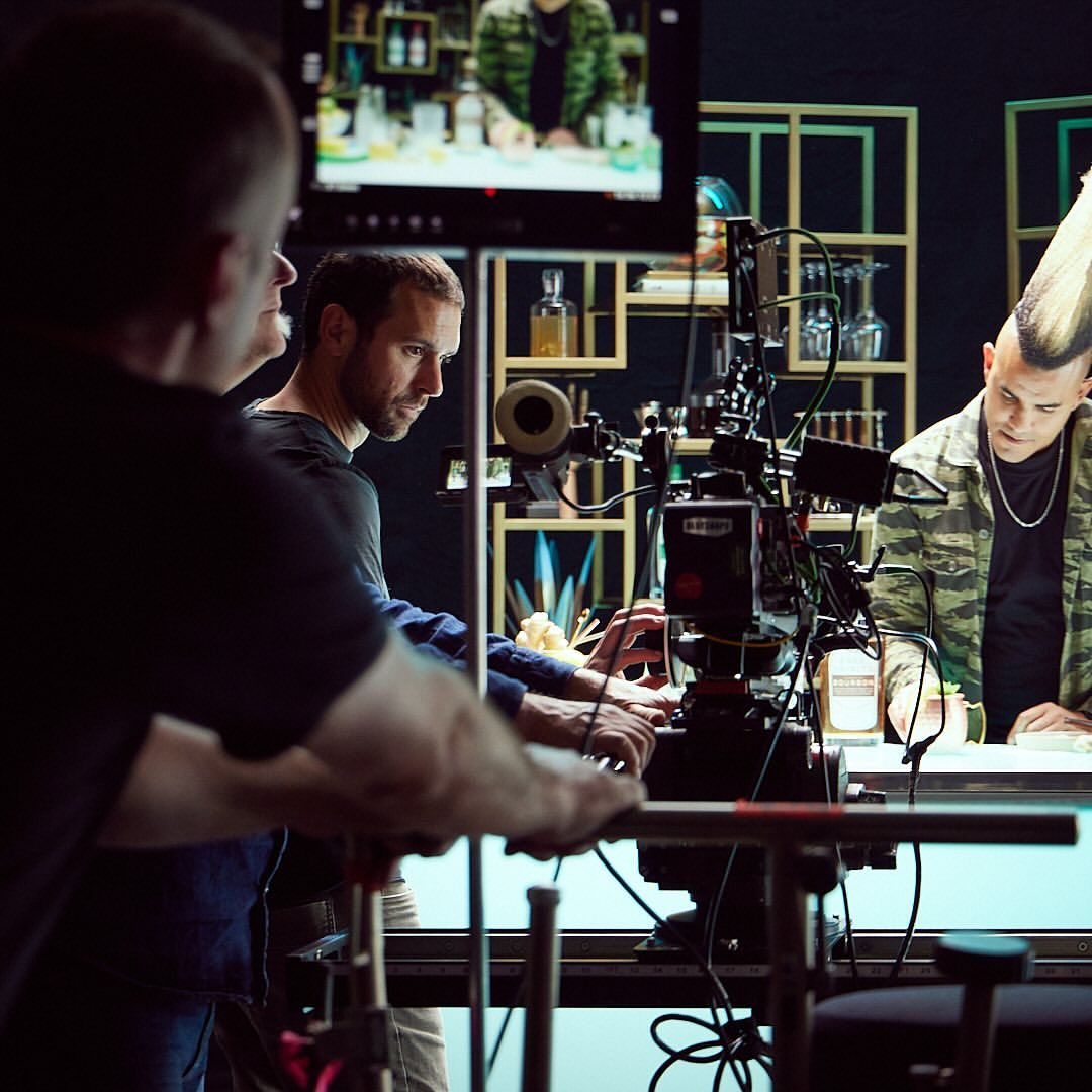 A behind-the-scenes shot of a film or television production, featuring crew members and an actor with a mohawk at a bar setup.