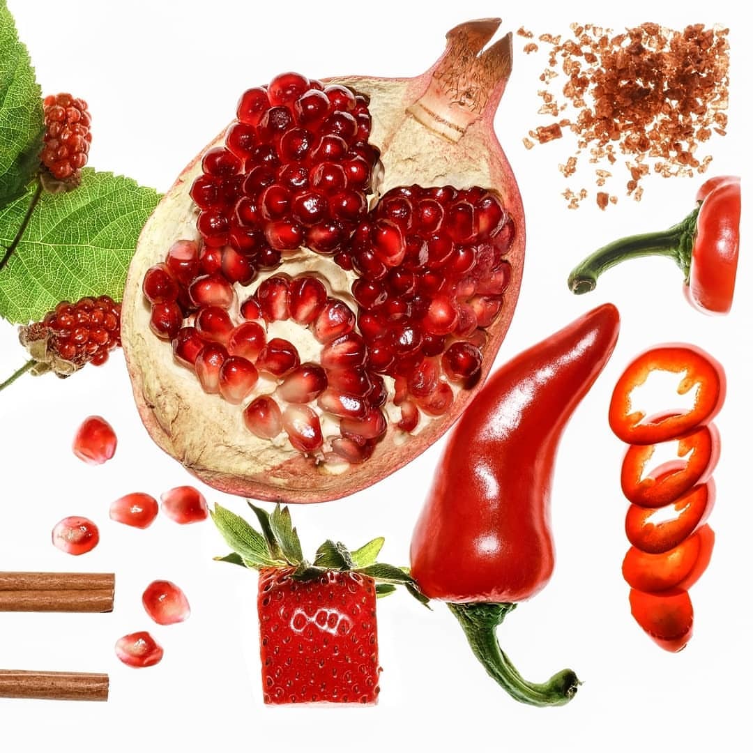Still life featuring a halved pomegranate, red chili pepper, cinnamon sticks, strawberry, and blackberries against a white background.
