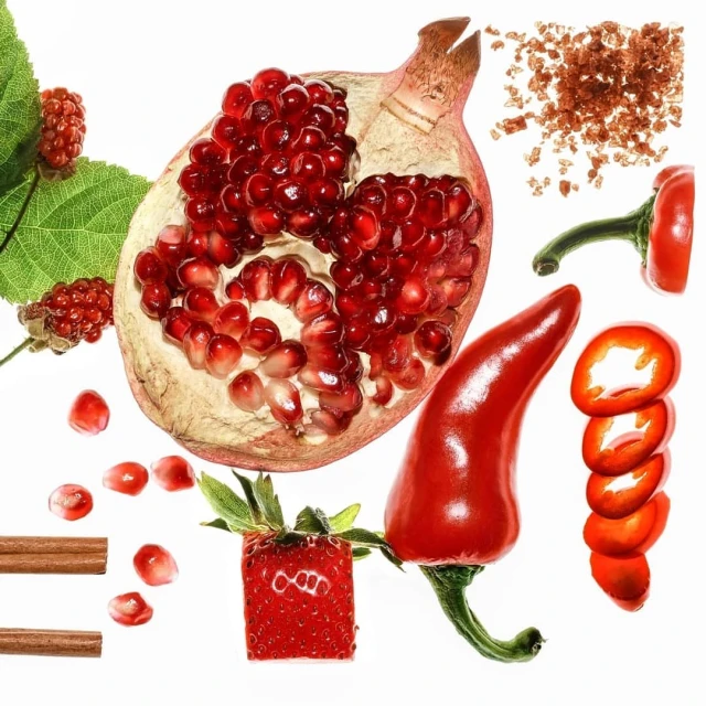 Still life featuring a halved pomegranate, red chili pepper, cinnamon sticks, strawberry, and blackberries against a white background.