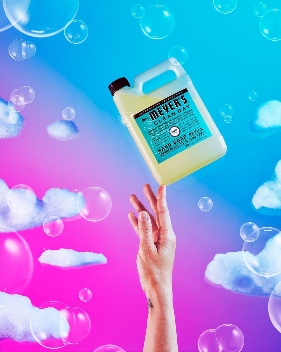Hand balancing bottle of Mrs. Meyer's Clean Day soap refill against a pink and blue background with cloud and bubble graphics.