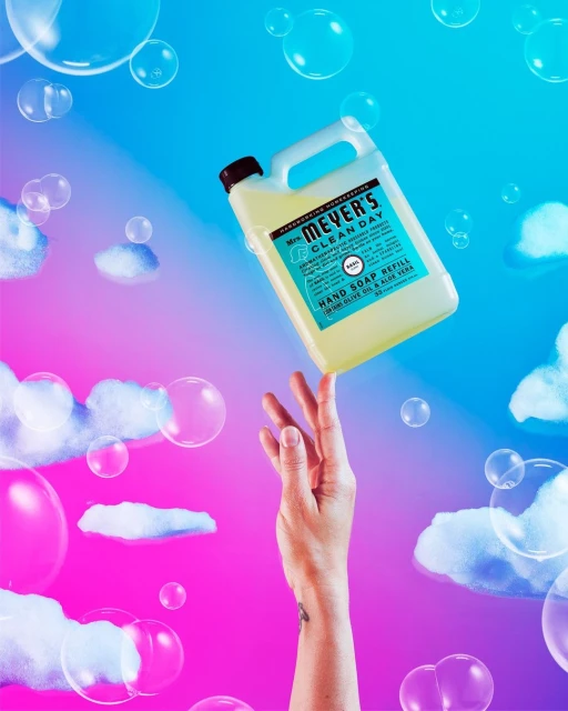 Hand balancing bottle of Mrs. Meyer's Clean Day soap refill against a pink and blue background with cloud and bubble graphics.