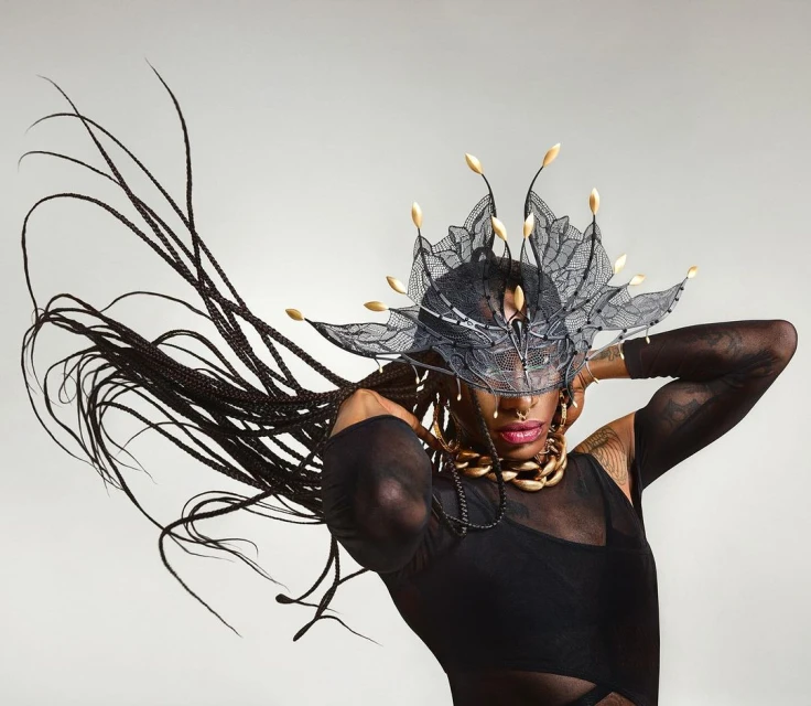 Woman in black bodysuit poses with elaborate black lace and gold headpiece and braided hair extensions against a neutral backdrop.