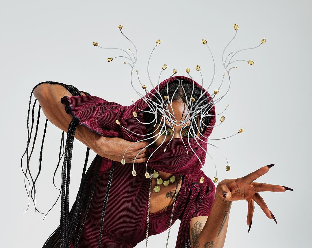 Portrait of an adult female wearing a maroon hooded dress and a silver metal headpiece, posing against a light grey studio backdrop.