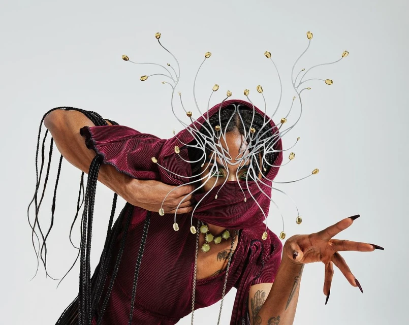 Portrait of an adult female wearing a maroon hooded dress and a silver metal headpiece, posing against a light grey studio backdrop.