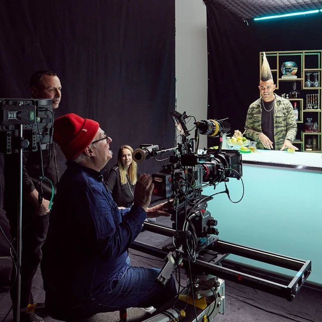 A film crew shoots a man with a mohawk at a bar, focusing on the camera and crew members in a studio setting with black backdrops.