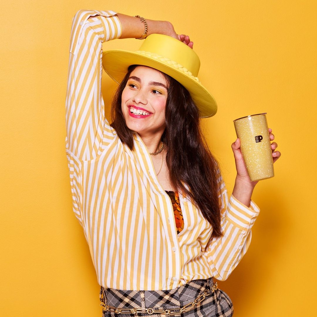 Young woman in striped shirt, hat, and plaid skirt, holding drink, smiles against a yellow backdrop in a vibrant, joyful studio shot.